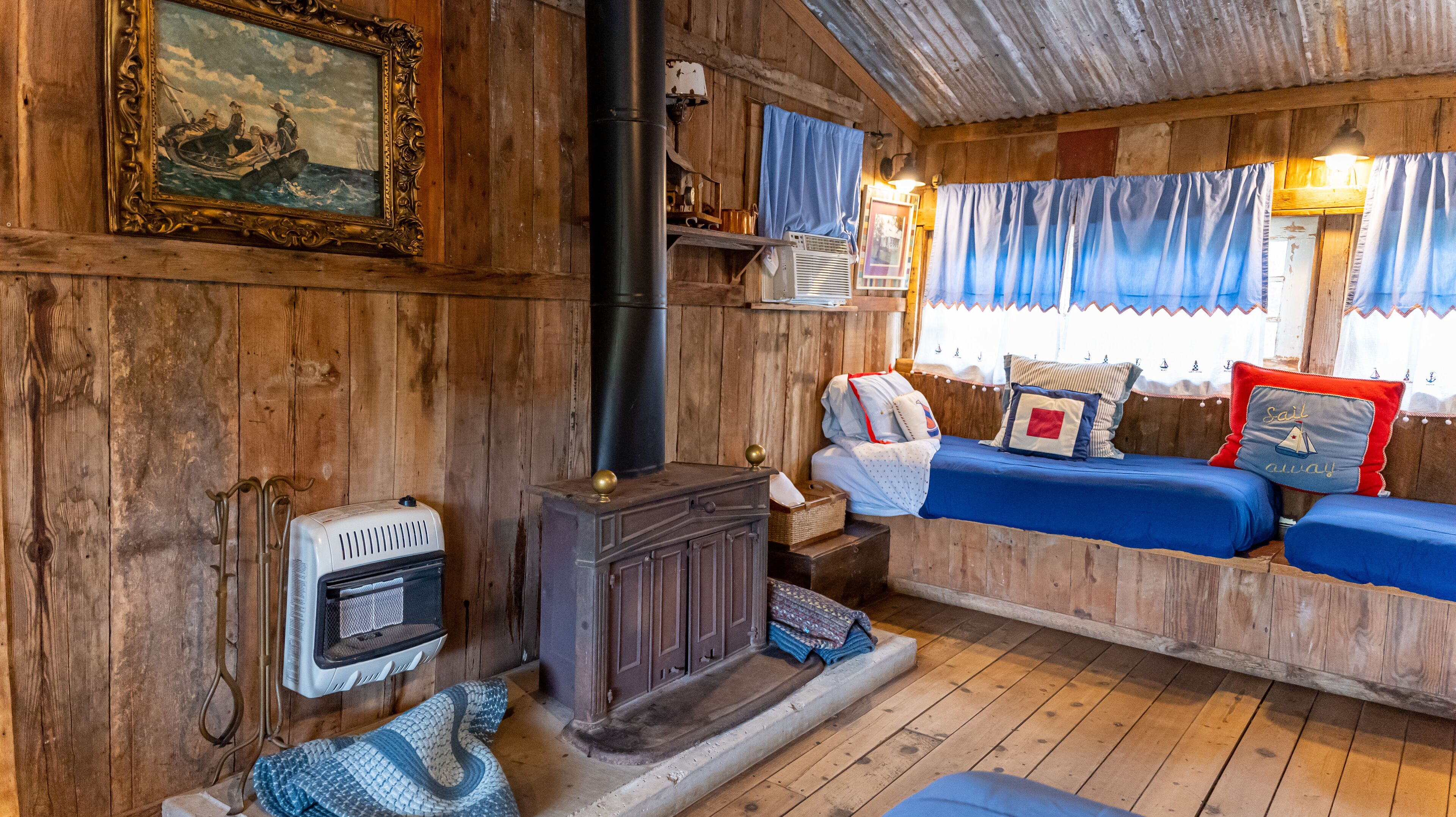 A cozy cabin interior with a wooden wall, a vintage stove, a bed with blue bedding, and a framed painting.
