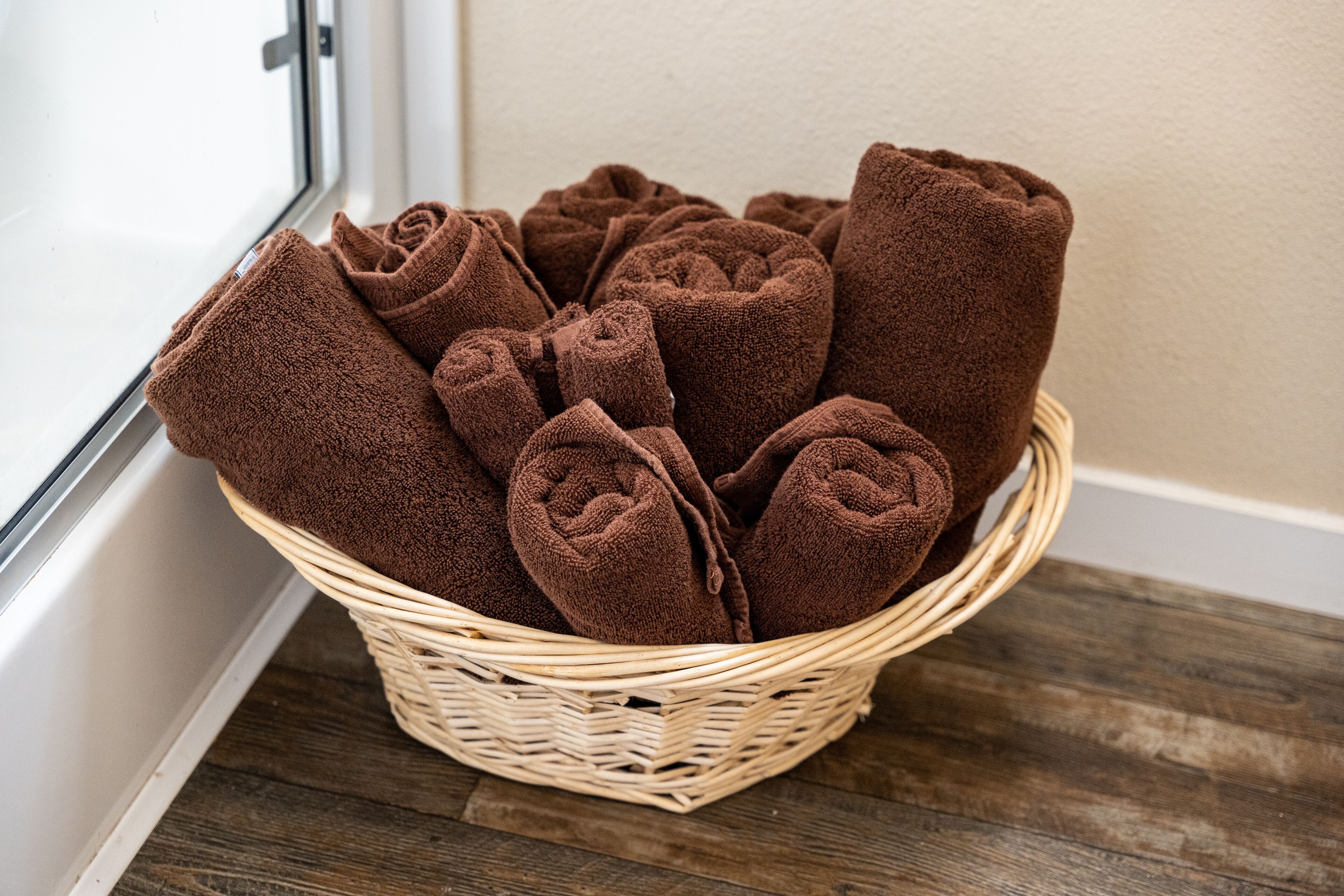 Een rieten mand gevuld met netjes opgerolde bruine handdoeken, geplaatst op een houten oppervlak nabij een raam.