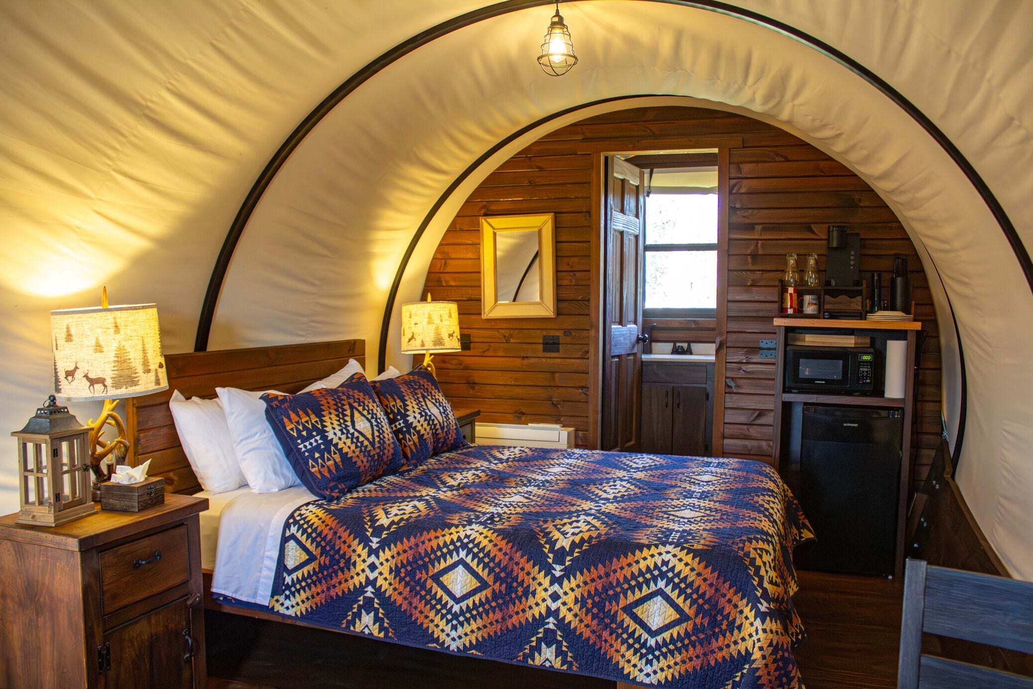 A tented accommodation with a bed, bedside lamps, a small kitchenette, and a wooden interior.