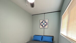 Une chambre à coucher comprenant un lit, un ventilateur de plafond, une tête de lit matelassée, une couvre-lit matelassée, un plancher en bois, une fenêtre avec des stores et une décoration murale matelassée.