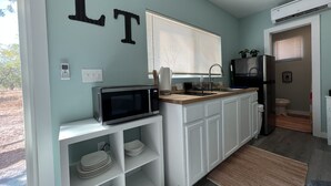 Une cuisine moderne avec des meubles blancs, un four à micro-ondes, un évier et un réfrigérateur. Il y a une fenêtre avec des stores et une porte qui donne sur une salle de bain.