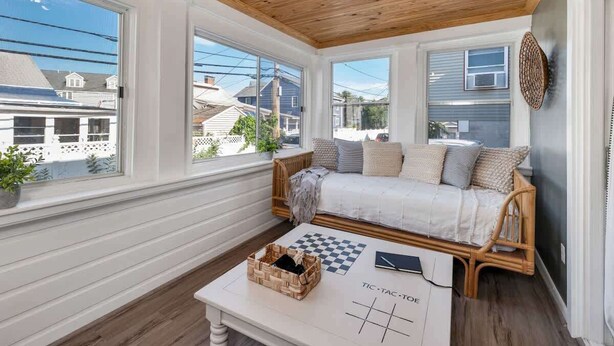 A cozy room with a wooden ceiling, a wicker sofa, a checkered table, and large windows offering a view of neighboring houses. - The Fleet -  Perfect Location Near Beach & Boardwalk (Hampton)