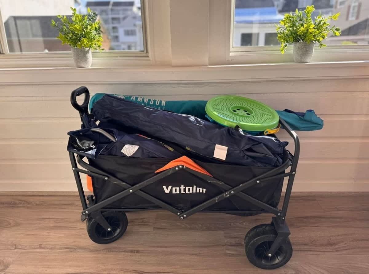 Een Votoim kinderwagen met een zwart frame en oranje accenten, op een houten vloer nabij een raam met twee planten in potten.