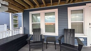 A covered porch with a wooden deck, two windows, and a black bench. - Riptide Retreat - Walk to Hampton & Seabrook Beach! (Seabrook)