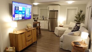 A living room with a TV, wooden furniture, a sofa, a small table, and a kitchen area with a fridge. - Riptide Retreat - Walk to Hampton & Seabrook Beach! (Seabrook)