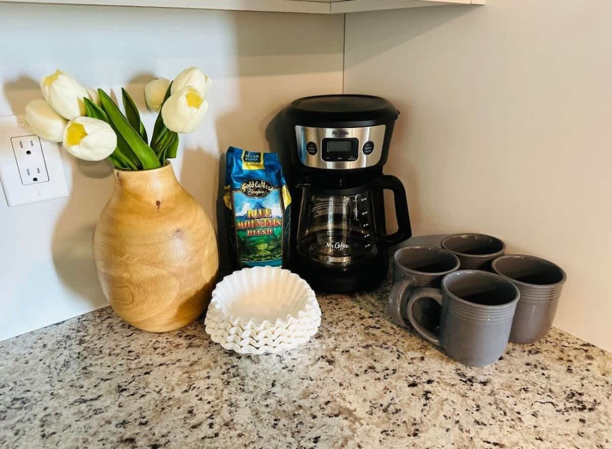 A kitchen counter with a coffee maker, a vase with tulips, a bag of coffee, and several mugs.