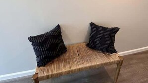 Un banco de madera con asiento y respaldo trenzados, dos almohadones oscuros y un adorno de madera fijado a la pared.