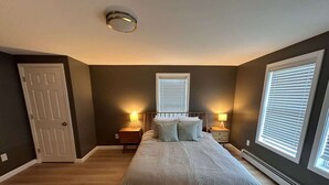Un dormitorio con una cama, dos mesitas de noche, una puerta y dos ventanas con persianas.