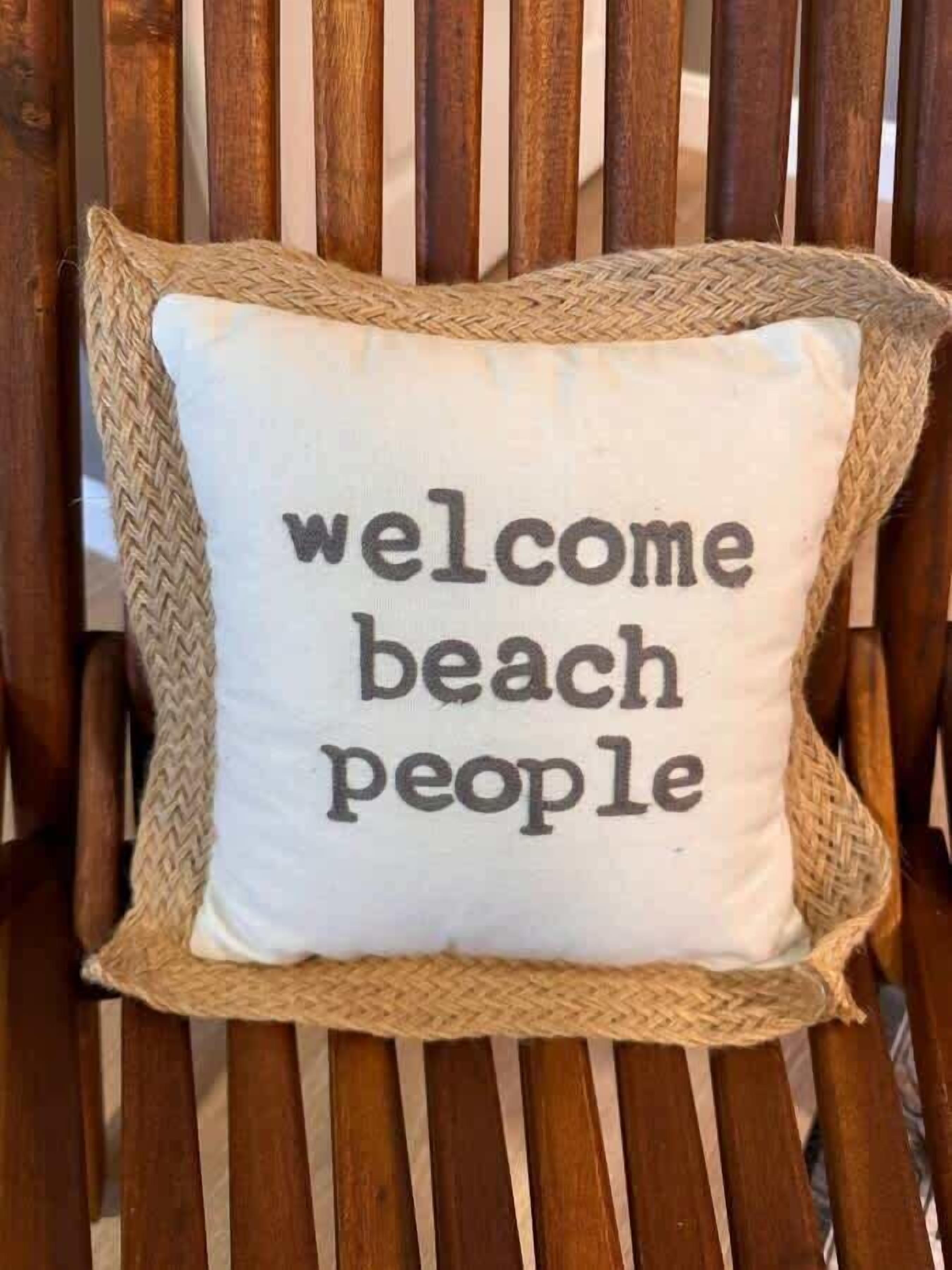 Un coussin blanc sur lequel est inscrit « bienvenue à tous, amateurs de plage », posé sur une chaise en bois.