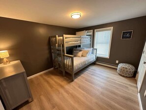 A bedroom with a bunk bed, a bedside table with a lamp, a window with blinds, and a wall-mounted sign.