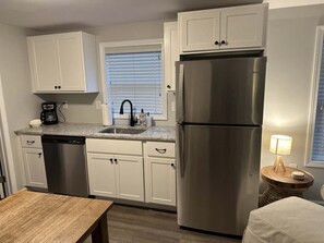 A compact kitchen with white cabinets, a stainless steel refrigerator, a dishwasher, a coffee maker, and a small round table with a lamp.