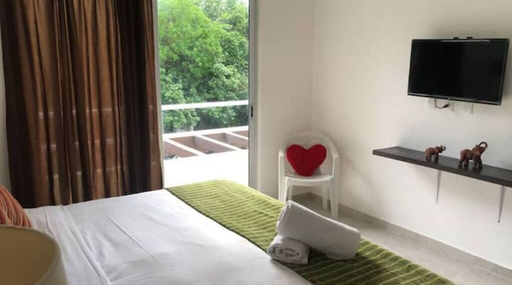 A hotel room with a bed, a green striped area rug, a white chair with a red heart cushion, a shelf with decorative items, and a TV mounted on the wall.