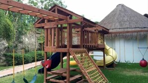 A wooden play structure with a slide and swings, set against a backdrop of a thatched-roof hut and lush greenery.