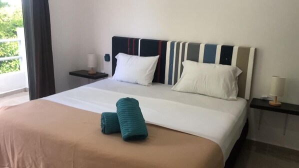 A hotel room with a bed, pillows, a nightstand, and a view of greenery outside.
