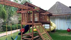 Una estructura de juego de madera con tobogán y columpios, frente a una zona verde con un edificio de techo de paja al fondo.