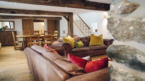 A cozy living room with a brown leather sofa, wooden dining table, and chairs. A staircase with a wooden railing is visible in the background.