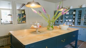 Una cocina con encimera de madera, un expositor de vidrio con queso, un florero con flores y un armario azul con puertas de cristal.