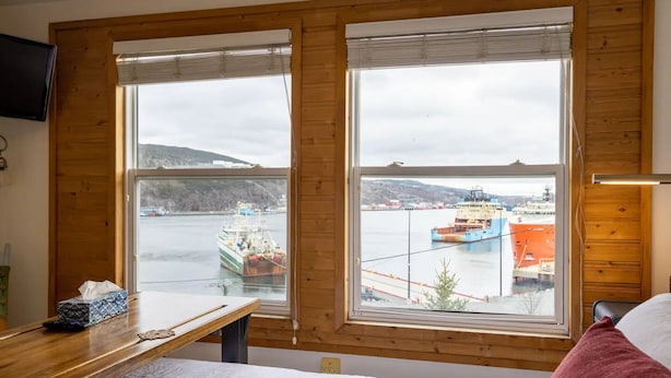 A room with a wooden desk, a bed, and large windows overlooking a harbor with boats. - Iconic Battery Cottage Signal Hill Sea&City Views (St. John's)
