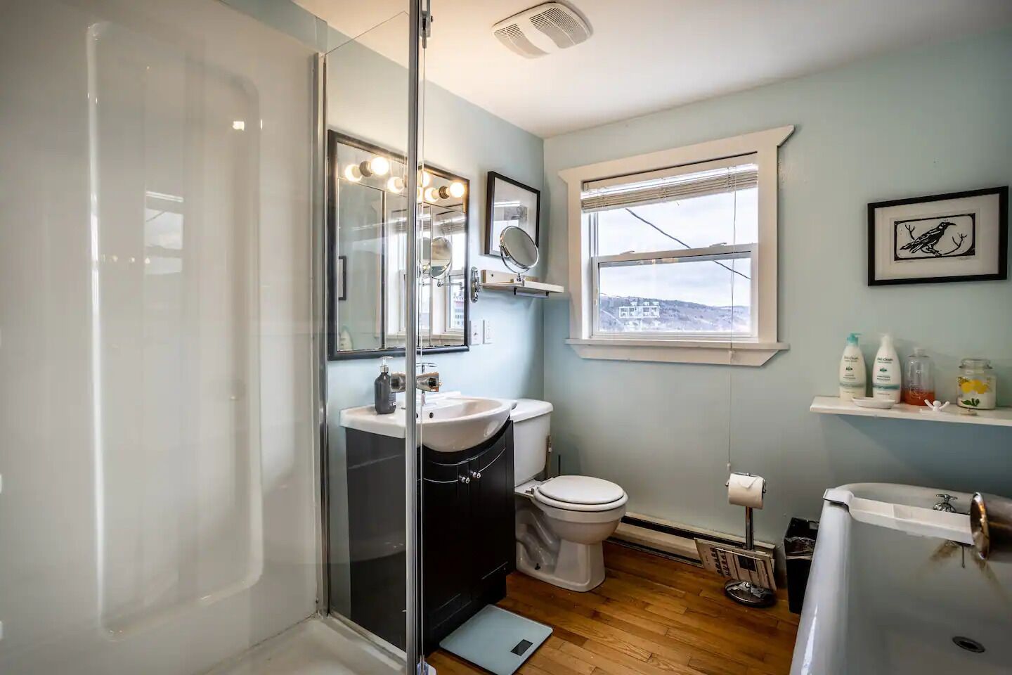 A bathroom with a glass shower enclosure, a toilet, a sink, and a window with a view of a building.