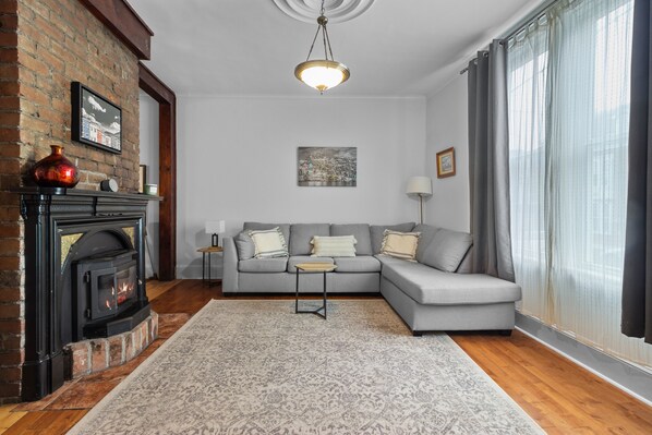 Una sala de estar con chimenea, un sofá gris, un tapete estampado y un ventanal con cortinas.