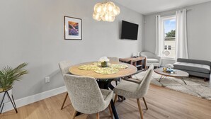 A modern living room with a round dining table, grey upholstered chairs, a flat-screen TV mounted on the wall, and a grey sofa.