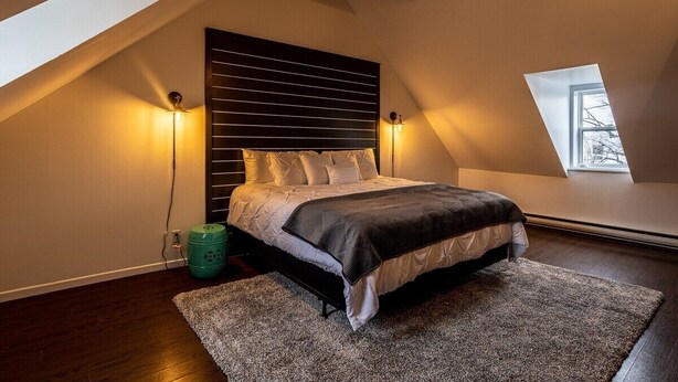A bedroom with a sloped ceiling, a bed with a headboard, two bedside lamps, a window, and a green trash bin. - The Bannerman Park Loft Private Parking King Bed (St. John's)