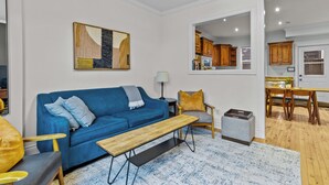 Sala de estar con un sofá azul, una mesa de centro de madera y un sillón gris. Hay un cuadro en la pared y se ve una cocina a través de una puerta abierta.
