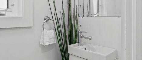 Une petite salle de bain blanche, avec un seul lavabo, un miroir et une fenêtre.