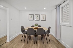 Uma sala de jantar com uma mesa de madeira, seis cadeiras e três plantas em vasos, emolduradas, na parede.