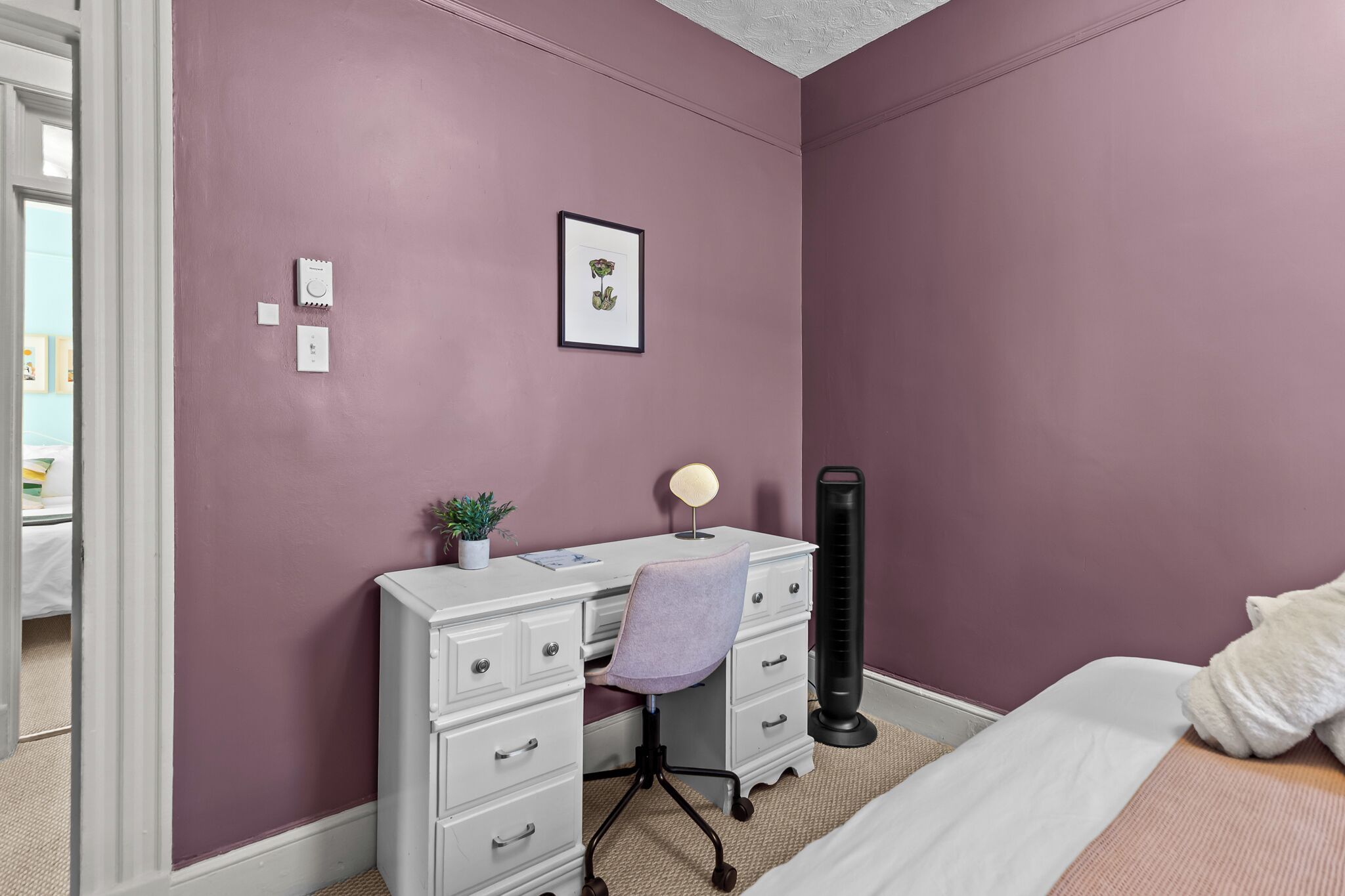 A bedroom with a bed, a desk with a chair, a standing heater, and a framed picture on the wall.