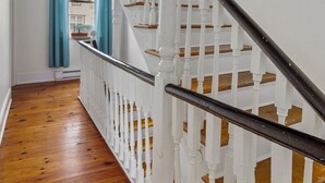 Un escalier avec des balustrades blanches et une main courante noire, menant à un palier d’où l’on peut voir une maison à travers une fenêtre ornée de rideaux bleus.