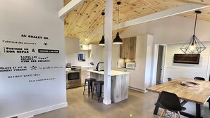 A modern kitchen with a central island, pendant lights, and a dining area with a wooden table and chairs. - A 30 Minutes de St-sauveur, Chalet Tout Équipé, Spa, Bord de lac (Gore)