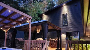 A wooden deck with a hot tub, a covered seating area, and a house with a dark exterior. - A 30 Minutes de St-sauveur, Chalet Tout Équipé, Spa, Bord de lac (Gore)