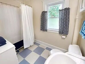 Ein Badezimmer mit Schachbrettboden, einer weißen Badewanne, einem weißen Waschbecken und einem Fenster mit gemusterten Vorhängen.