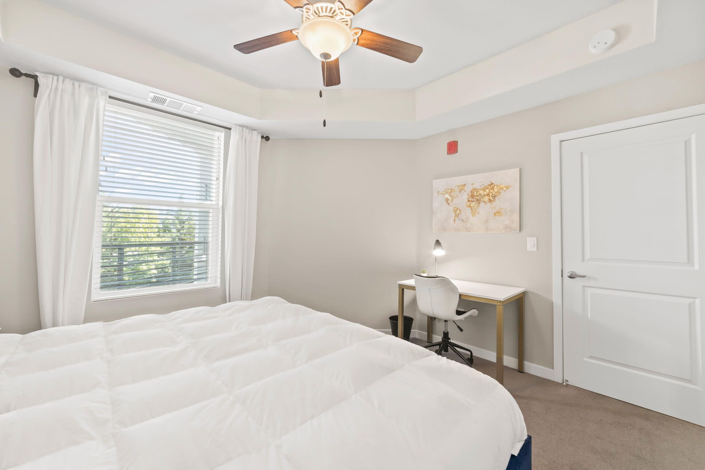 Une chambre à coucher avec un grand lit, un bureau avec une chaise, un ventilateur de plafond et une fenêtre avec des stores.