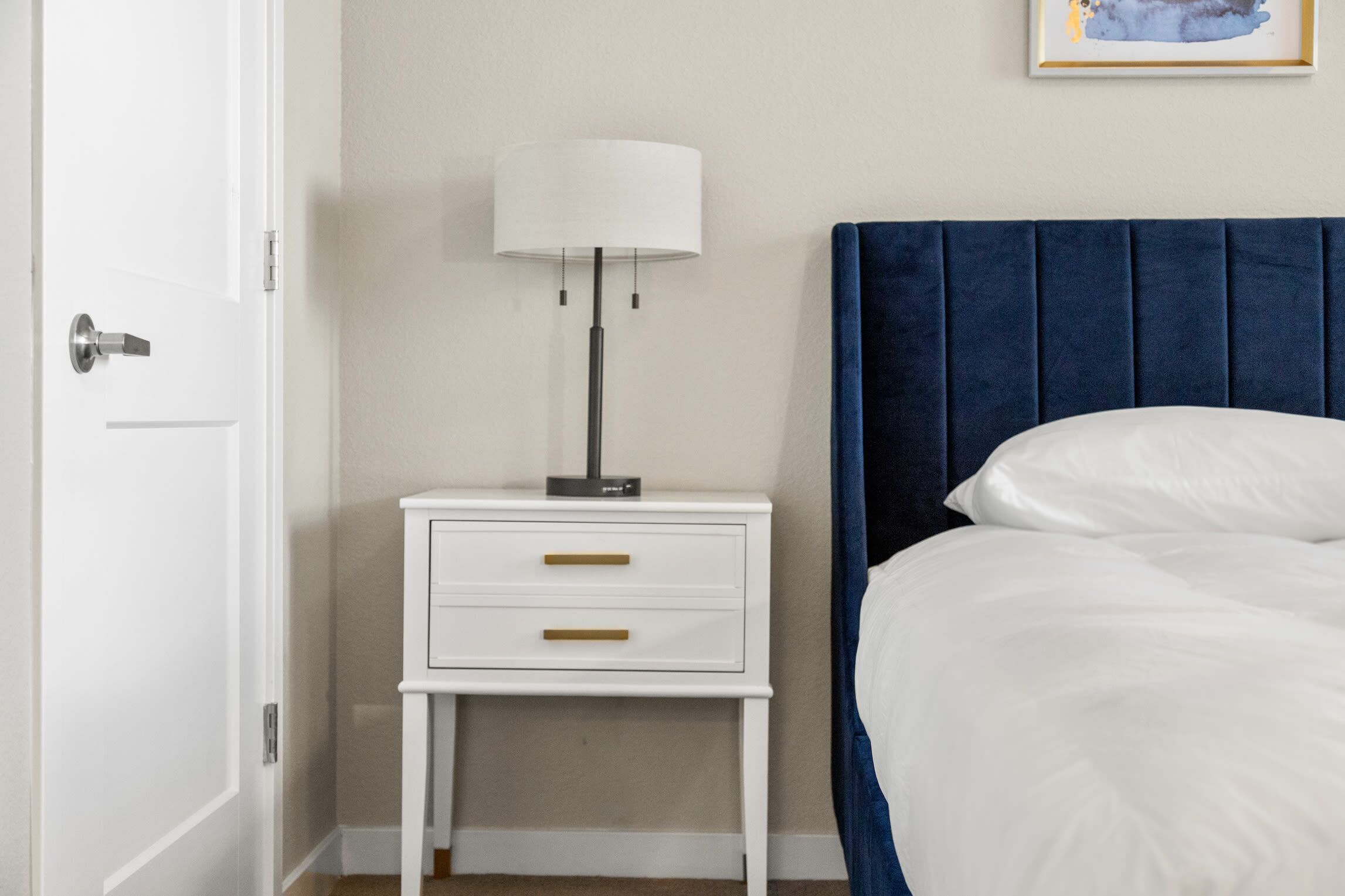 A bedroom with a white nightstand, a lamp, a blue headboard, and a white bed.