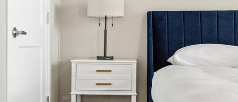 A bedroom with a white nightstand, a lamp, a blue headboard, and a white bed.