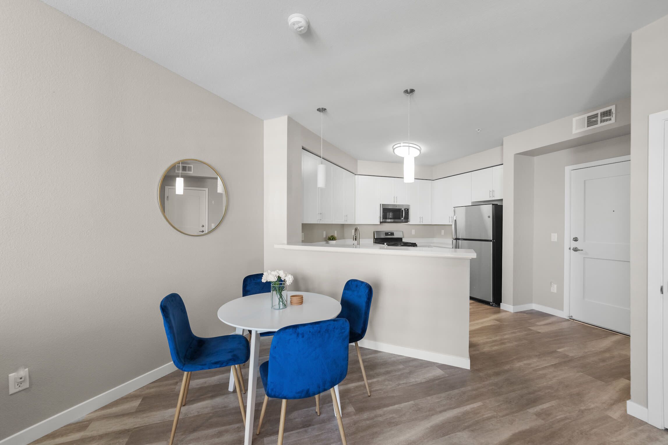 A modern kitchen with white cabinets, a central island, and stainless steel appliances. There is a dining area with a round table and blue chairs.