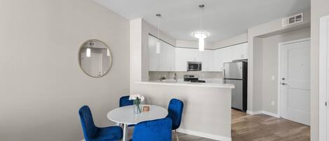 A modern kitchen with white cabinets, a central island, and stainless steel appliances. There is a dining area with a round table and blue chairs.