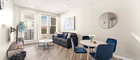 A modern living room with a sofa, a dining table with chairs, a round mirror, and a small side table with a vase and a plant.