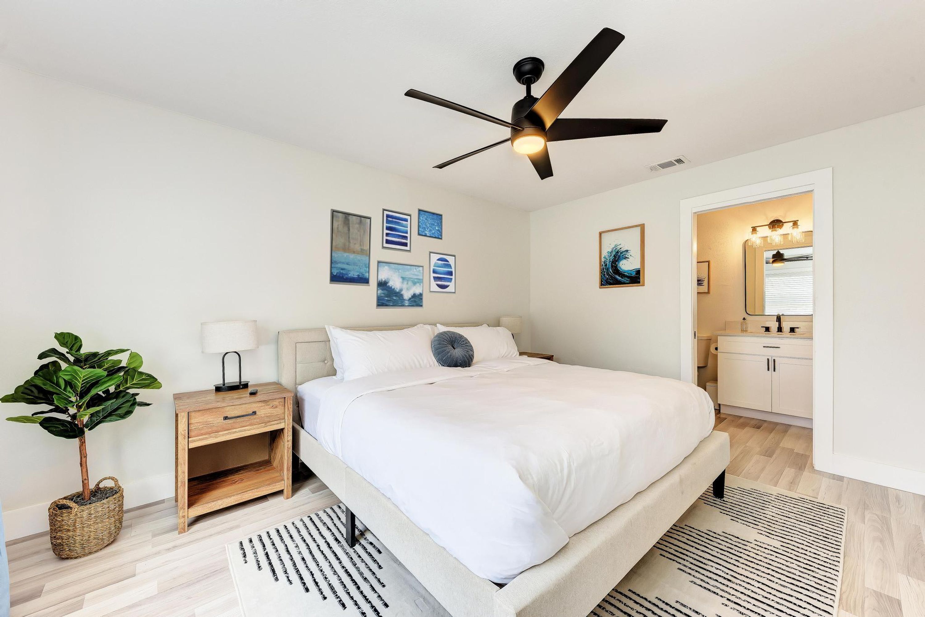 A bedroom with a bed, bedside table, lamp, ceiling fan, wall art, and a potted plant.