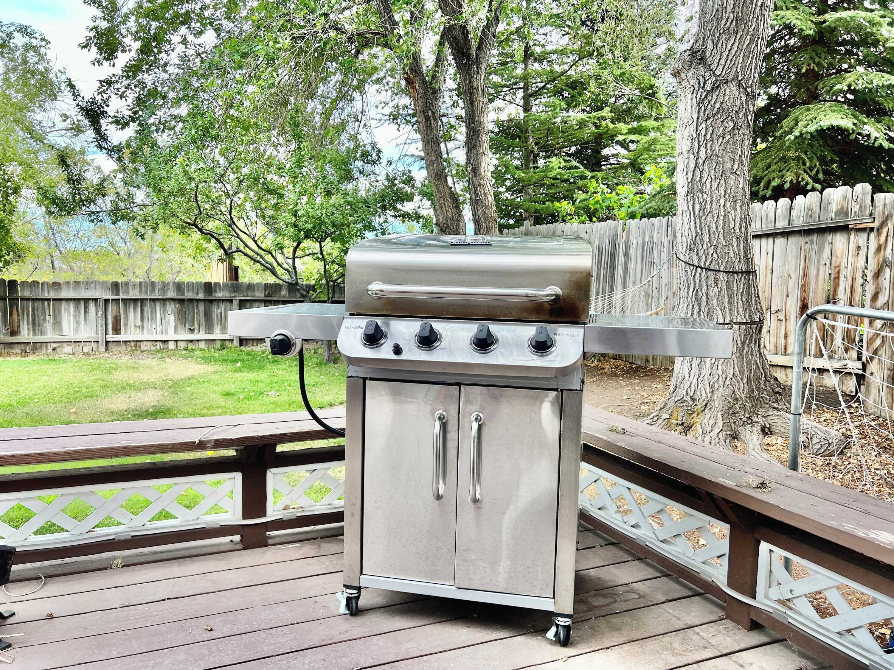 Una griglia per barbecue in acciaio inox con quattro fuochi, un pannello di controllo e un mobiletto portaoggetti, posizionata su un muretto in legno con una recinzione in legno sullo sfondo.