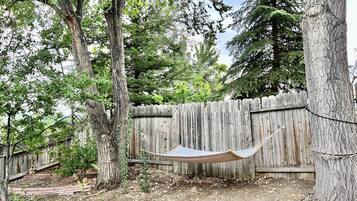 Una hamaca entre árboles en un patio trasero con valla de madera y un sendero de ladrillos.
