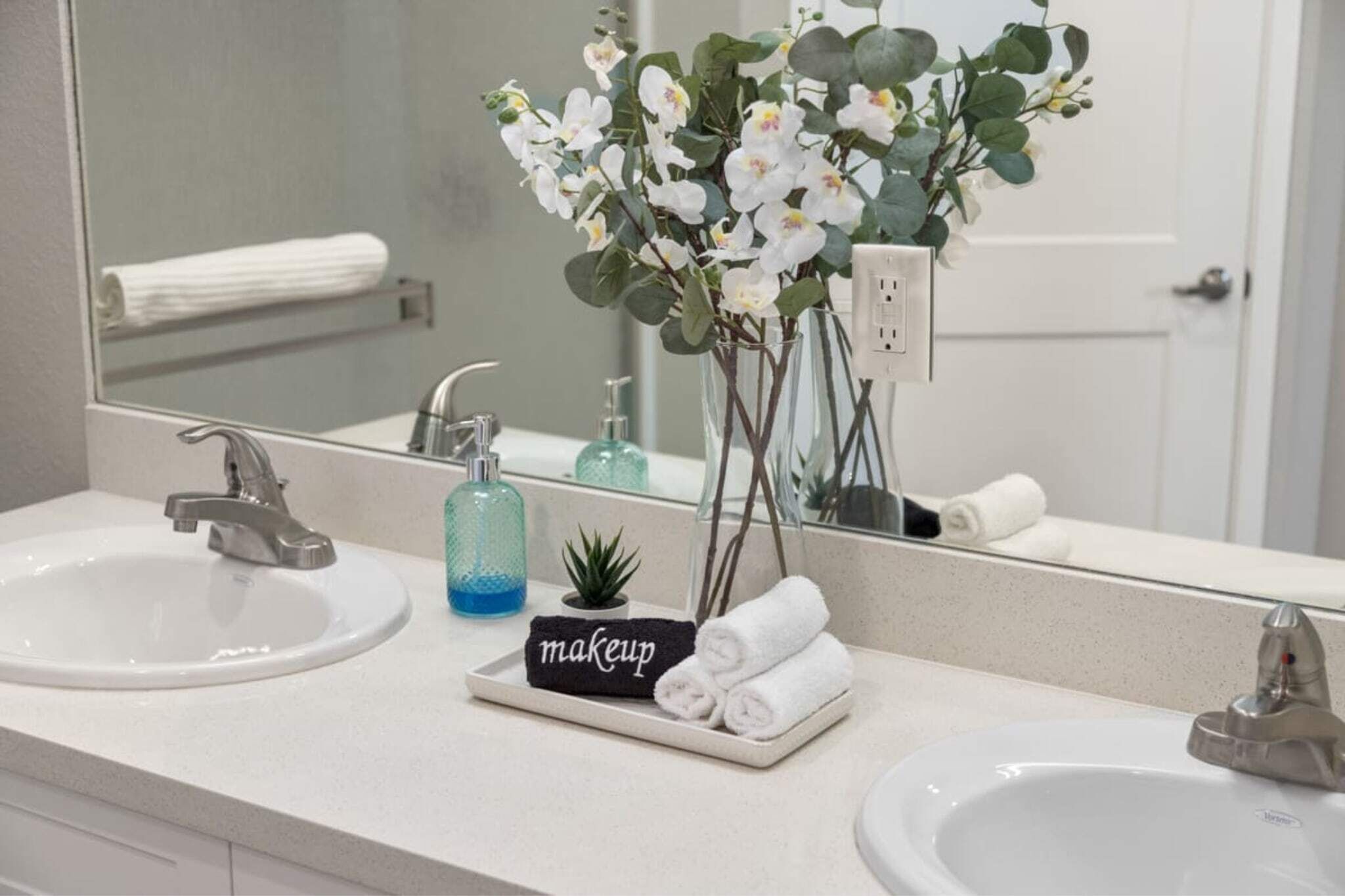 Un bagno con due lavandini, un ampio specchio e un vaso con fiori bianchi.