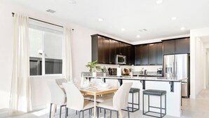 A modern kitchen with dark cabinets, a stainless steel refrigerator, and a dining area with white chairs and a wooden table. - Stylish Sleek 4BR Nevada Home Near Downtown Reno (Sparks)