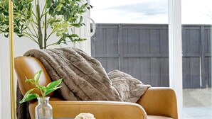 A cozy living room with a brown leather sofa, a blue round table with a vase and a macrame piece, and a view of a wooden fence outside. - Stylish Sleek 4BR Nevada Home Near Downtown Reno (Sparks)