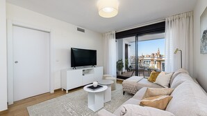 Una sala de estar moderna con un televisor de pantalla plana, muebles blancos, una mesita redonda y un ventanal con vistas a la ciudad.