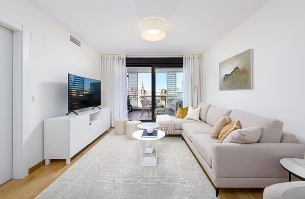 A modern living room with a flat-screen TV, a beige sofa, a small white coffee table, and a view of the city through large windows.