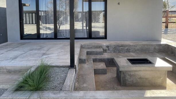 A modern outdoor patio area with a concrete floor, a pergola with a woven shade, and a large glass door leading to a deck. - MILU-CABAÑA SENCILLA (Guadalupe)