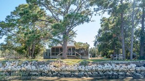 A cabin with a stone wall and a wooden deck, surrounded by trees and a body of water.